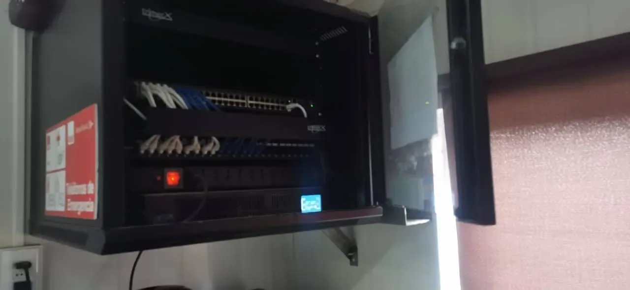 Gabinete de red abierto en un pasillo, con switch, cableado y cartel de emergencia